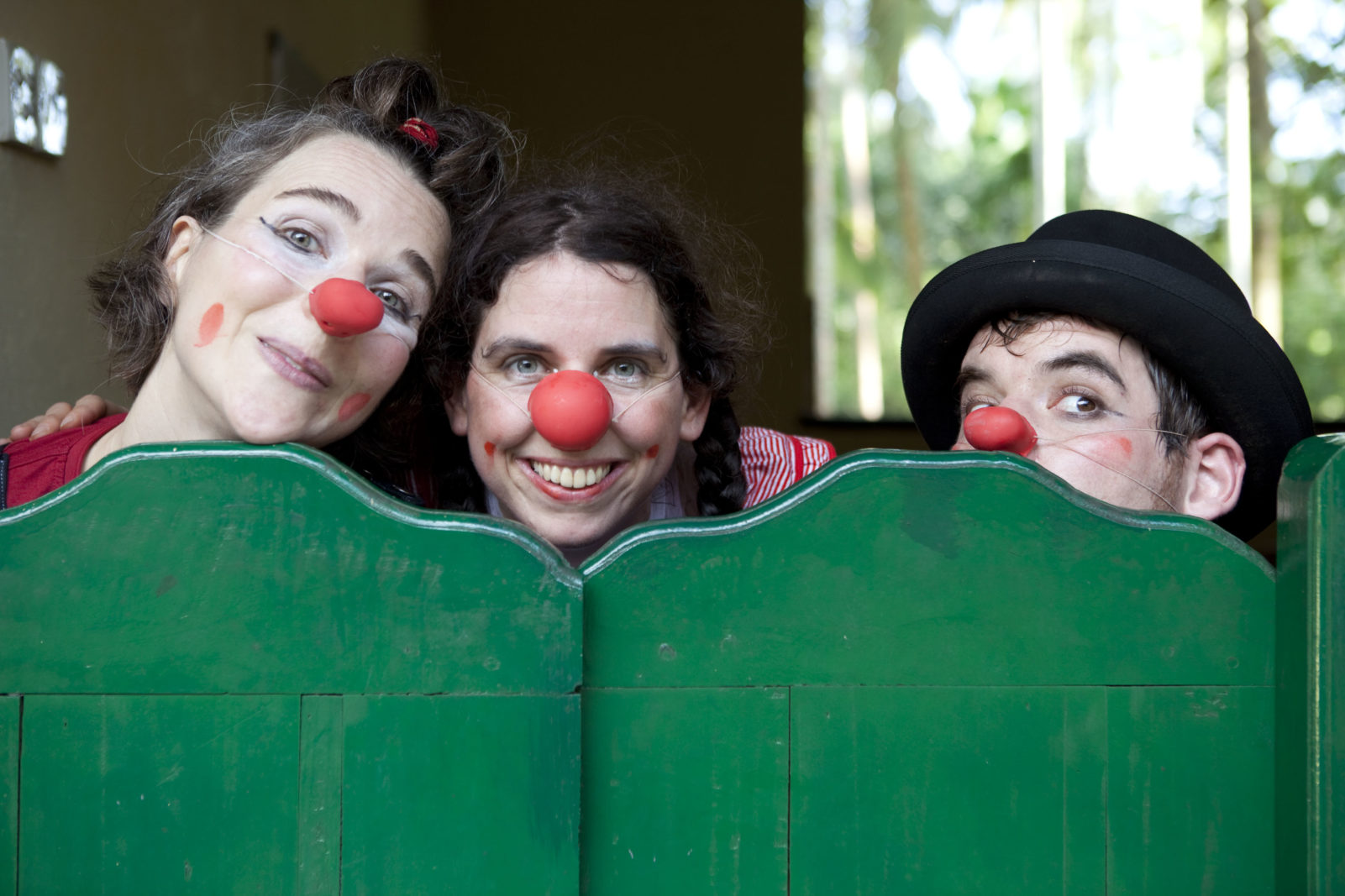 2011 Sri Lanka - Thomas Victor Drei Clowns mit roten Nasen und Gesichtsbemalung lächeln spielerisch, während sie über eine grüne Holzbarriere spähen. Ein Clown trägt einen schwarzen Hut. Im Hintergrund sind Sonnenlicht und Grünpflanzen zu sehen.
