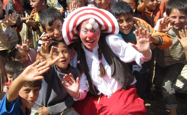 MITMACHEN_Syrien2013Mai_IMG_2627_ThomasVictorH374 Eine fröhliche Person mit einem rot-weißen Hut posiert mit einer Gruppe lächelnder Kinder im Freien, die alle aufgeregt mit den Händen in Richtung Kamera winken.