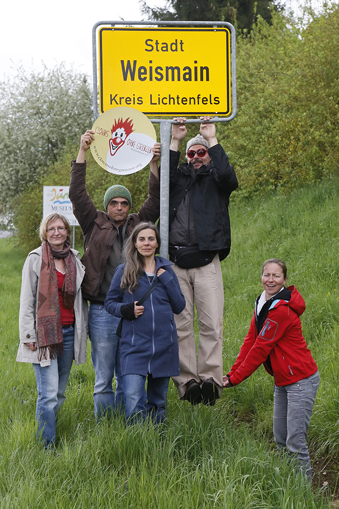 Weismain_01 Fünf Clowns posieren und lächeln neben einem gelben Ortsschild mit der Aufschrift Stadt Weismain Kreis Lichtenfels. Ein Clown hält ein rundes Schild, ein anderer hängt an der Stange. Sie stehen auf einer Wiese am Straßenrand.