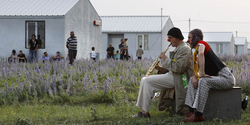 2010 Georgien - Manfred Lehner (3) - 800x400 Zwei Clowns sitzen auf einem Betonblock vor bescheidenen Häusern und einem Feld mit lila Blumen, einer spielt Saxophon. Mehrere Personen, darunter auch Kinder, schauen zu oder stehen im Hintergrund. Umgeschrieben mit Ihrer Anweisung: Zwei seltsam gekleidete oder rotnasige Personen (Clowns) sitzen auf einem Betonblock vor bescheidenen Häusern und einem lila Blumenfeld, einer spielt Saxophon. Mehrere Personen, darunter auch Kinder, schauen zu oder stehen im Hintergrund.