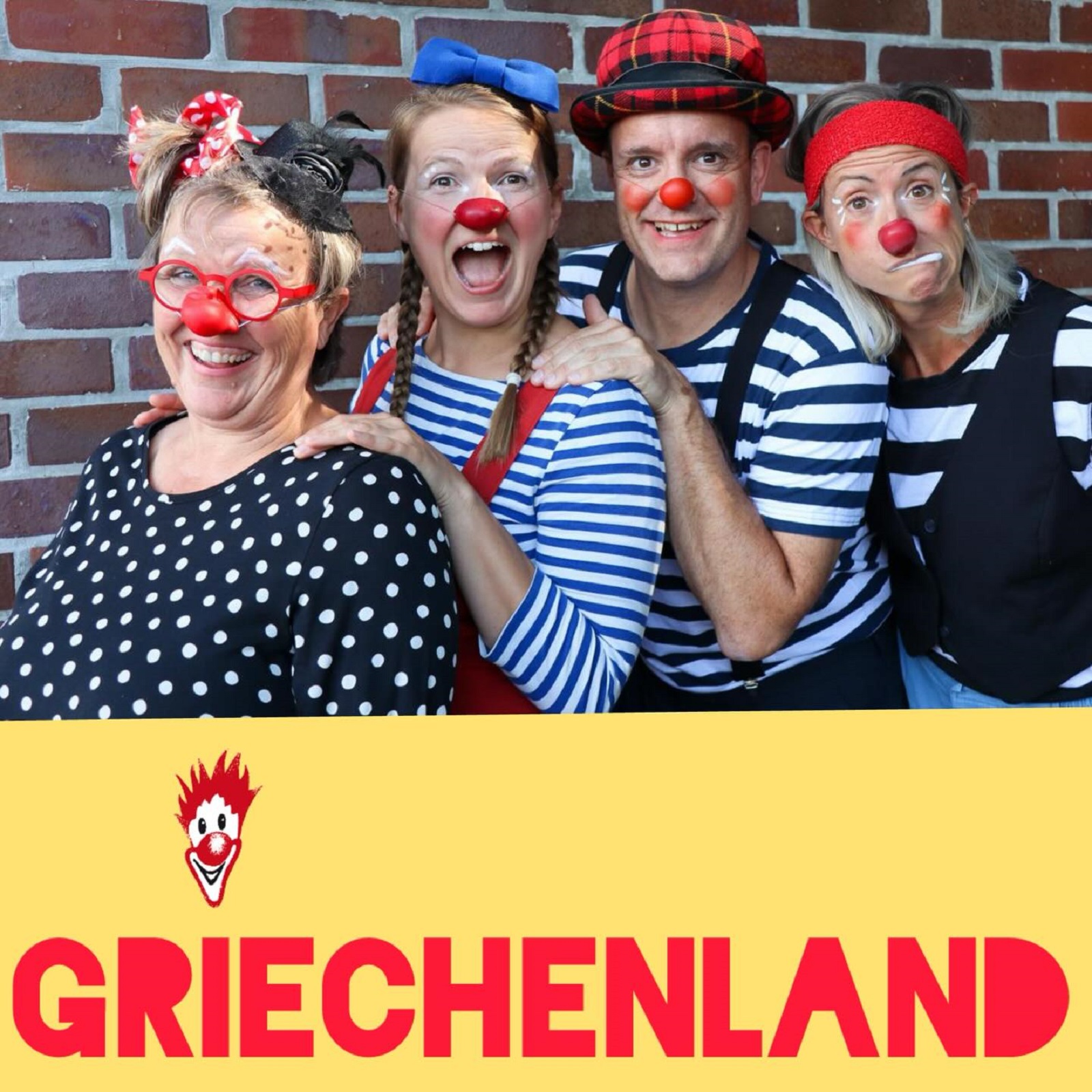 Vier Erwachsene mit roten Nasen, bunten Kostümen und einem breiten Grinsen stehen als Clowns verkleidet vor einer Backsteinmauer. Unter ihnen hängt ein gelbes Transparent mit einem Clownsgesicht und dem Wort GRIECHENLAND in fetten roten Buchstaben.