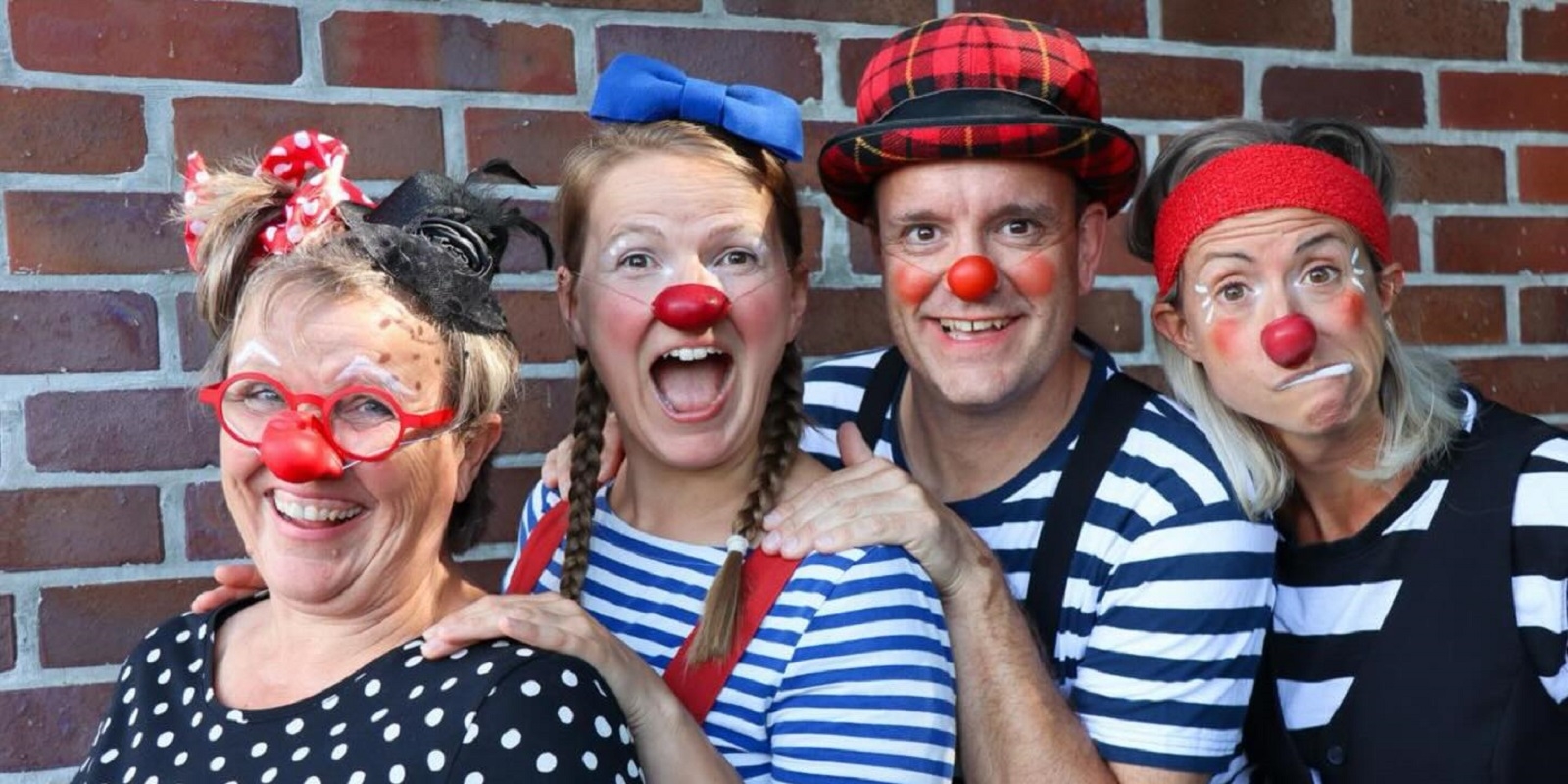 Vier Erwachsene, die als Clowns mit roten Nasen gekleidet sind, stehen vor einer Backsteinmauer, lächeln und machen lustige Grimassen. Sie tragen bunte Kostüme, Hüte und Accessoires wie Schleifen, Hosenträger und Tupfen.