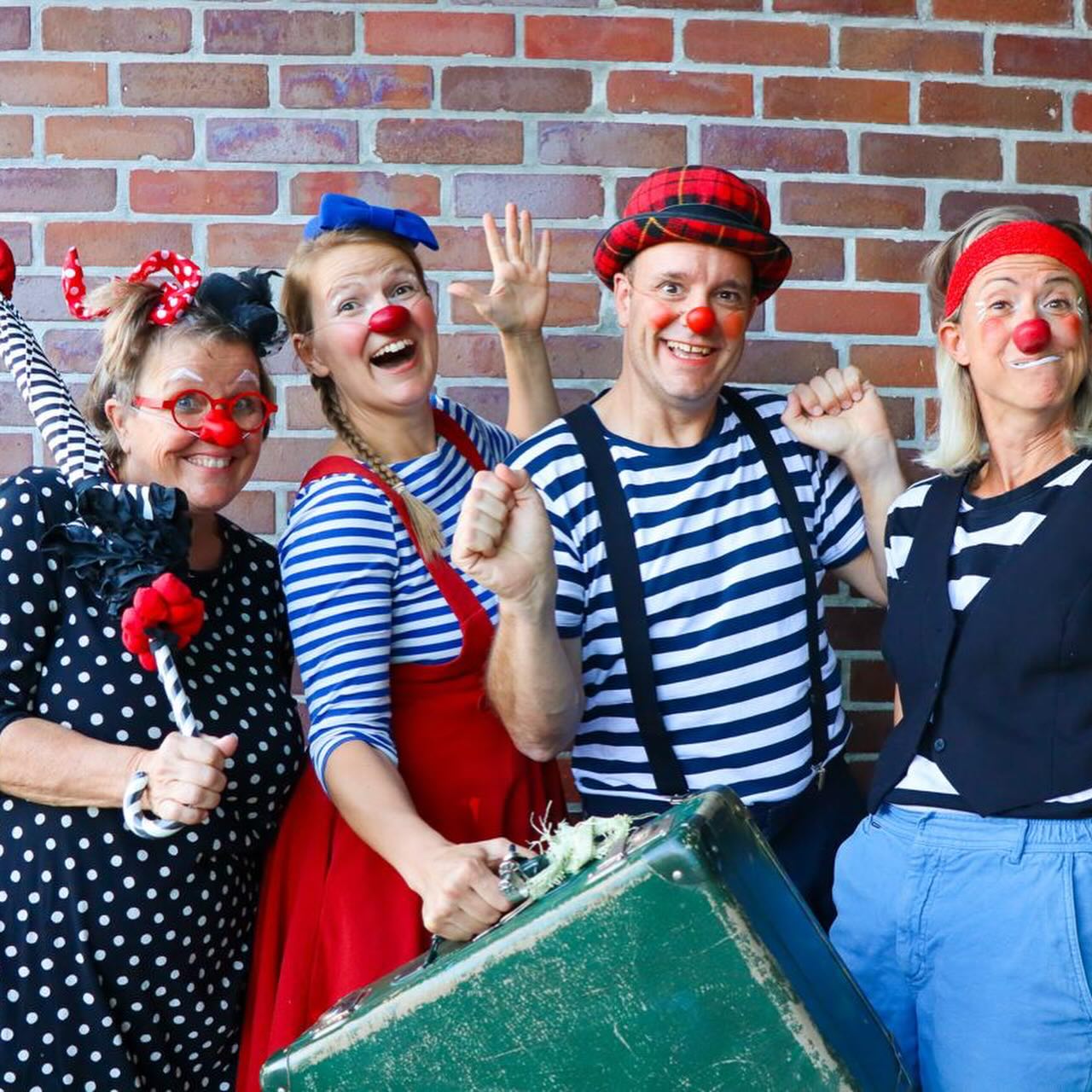 Vier Erwachsene, die als fröhliche Clowns mit roten Nasen und farbenfrohen Kostümen gekleidet sind, posieren gemeinsam vor einer Backsteinmauer, lächeln und winken; einer hält einen gestreiften Regenschirm, ein anderer einen grünen Koffer.
