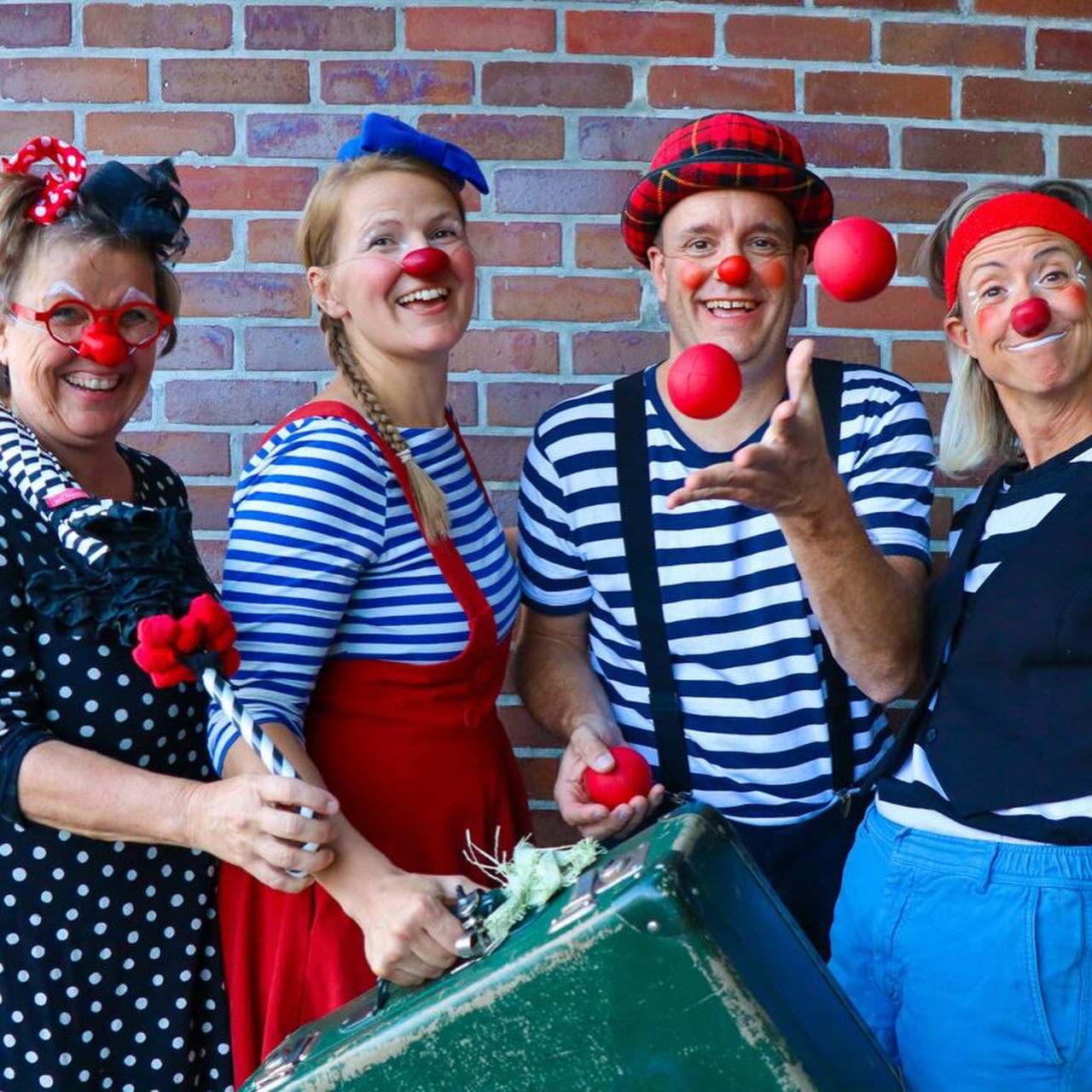 Vier als Clowns verkleidete Personen posieren mit roten Nasen und bunten Kostümen vor einer Backsteinmauer. Einer jongliert mit roten Bällen, ein anderer hält eine schwarz-weiße Requisite. Alle lächeln und sind fröhlich.