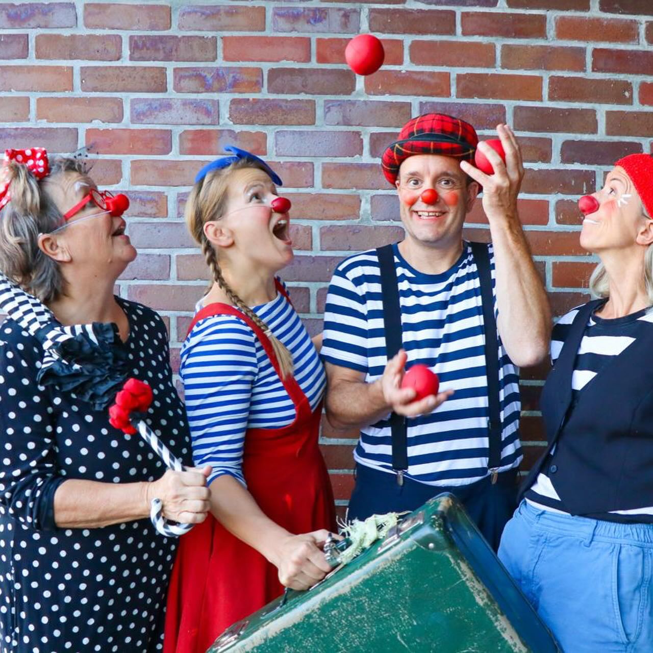 Vier Erwachsene, die als Clowns mit roten Nasen verkleidet sind, stehen vor einer Backsteinmauer. Einer jongliert mit roten Bällen, die anderen schauen zu und lächeln. Sie tragen verspielte, bunte Kostüme und Accessoires.