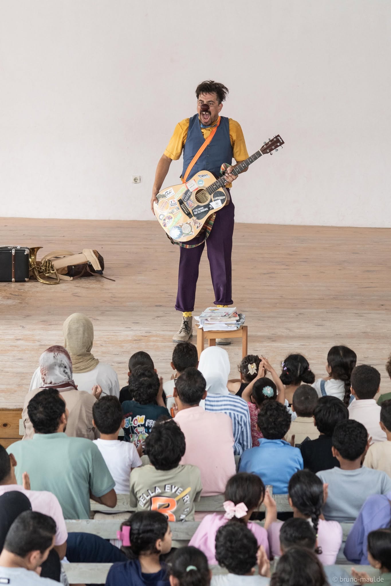 Ein Künstler mit einer großen, mit Aufklebern übersäten Gitarre unterhält ein sitzendes Publikum aus Kindern und Erwachsenen in einem hellen Innenraum. Die Zuschauer blicken zur Bühne und beobachten aufmerksam, wie er lebhafte Mimik zeigt.