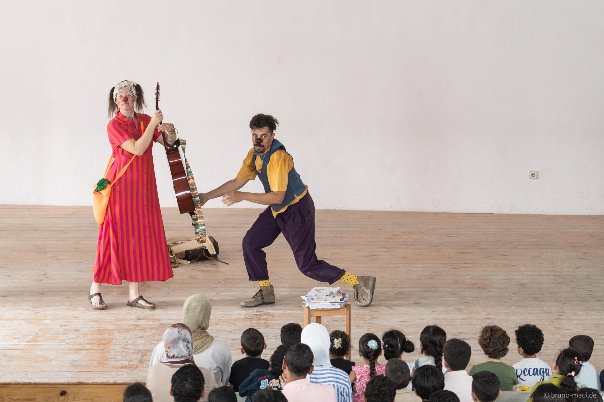 Zwei Clowns treten auf einer schlichten Holzbühne vor einem sitzenden Kinderpublikum auf. Ein Clown hält eine Gitarre in der Hand, während der andere eine verspielte Pose einnimmt. Die Kinder schauen aufmerksam vom Boden aus zu.