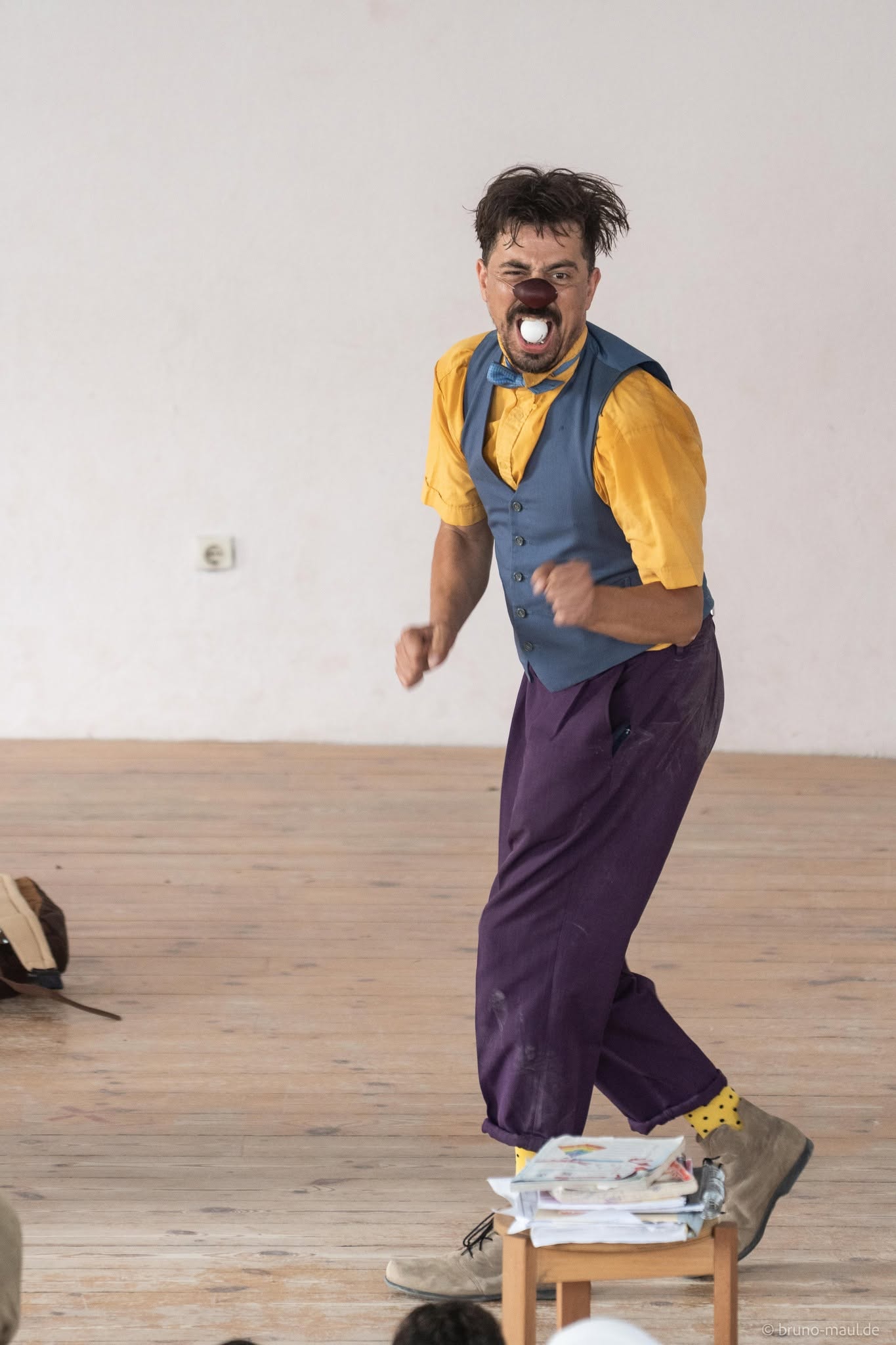 Ein Clown mit roter Nase, gelbem Hemd, blauer Weste und lila Hose tritt lebhaft auf einem Holzboden auf; auf einem kleinen Tisch liegen Papiere, und neben ihm auf dem Boden steht ein Hut.
