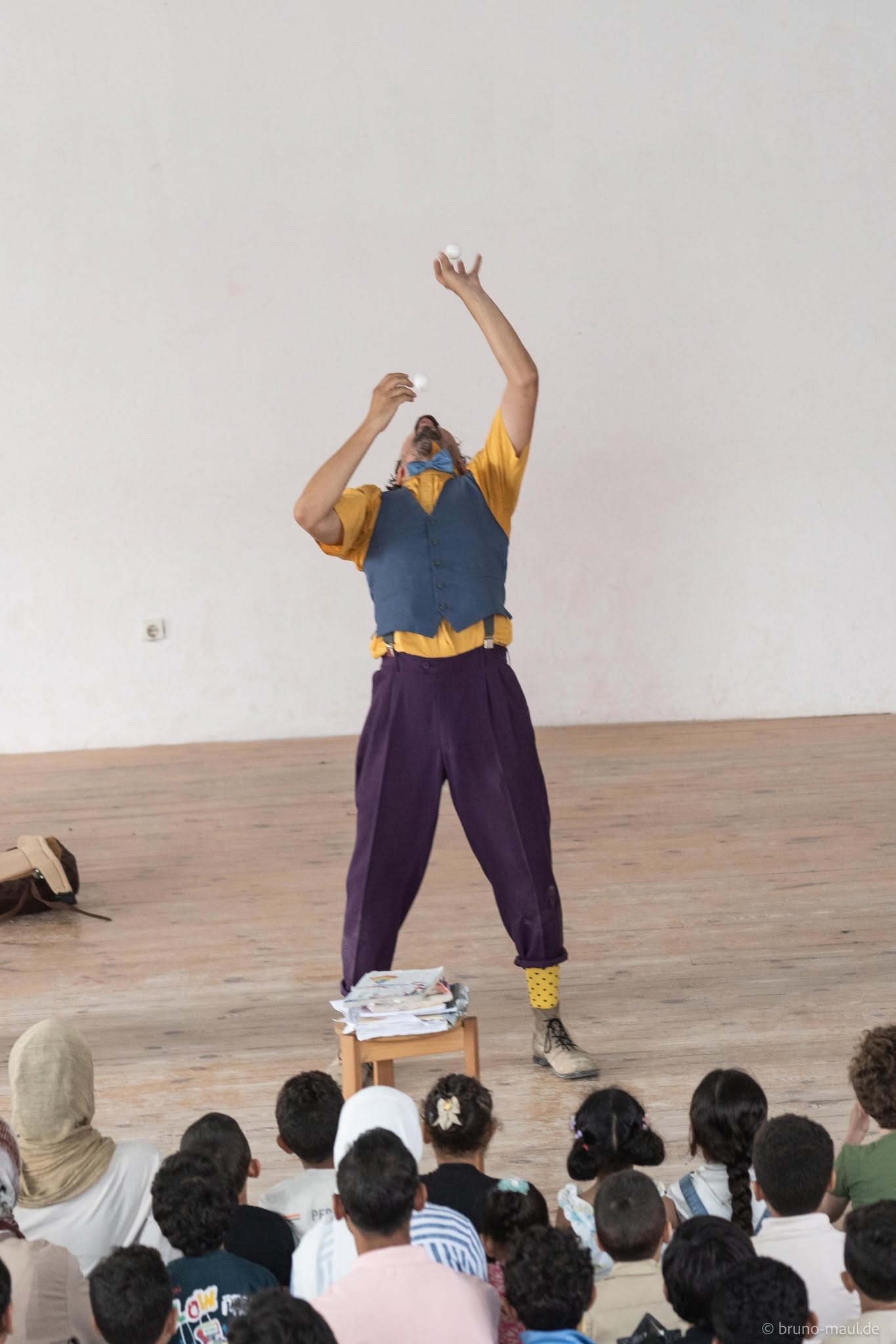 Ein als Clown verkleideter Künstler, der ein gelbes Hemd, eine blaue Weste und eine lila Hose trägt, jongliert, während er auf dem Rücken balanciert, und unterhält ein sitzendes Publikum aus Kindern und Erwachsenen in einem geräumigen Raum mit nacktem Boden und weißen Wänden.