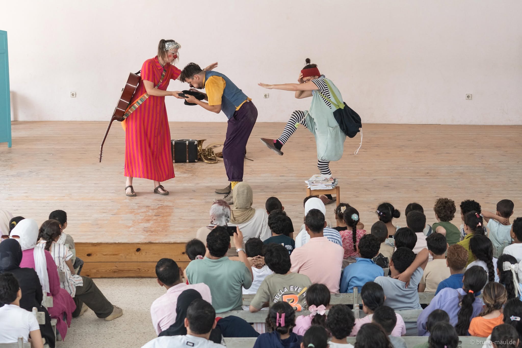 Drei Darsteller unterhalten eine sitzende Gruppe von Kindern in einer Turnhalle oder einem Saal. Eine trägt ein rotes Kleid und hat eine Gitarre dabei, eine andere beugt sich nach vorne, und eine dritte in gestreiften Strumpfhosen balanciert auf einem Hocker. Das Publikum schaut gebannt zu.