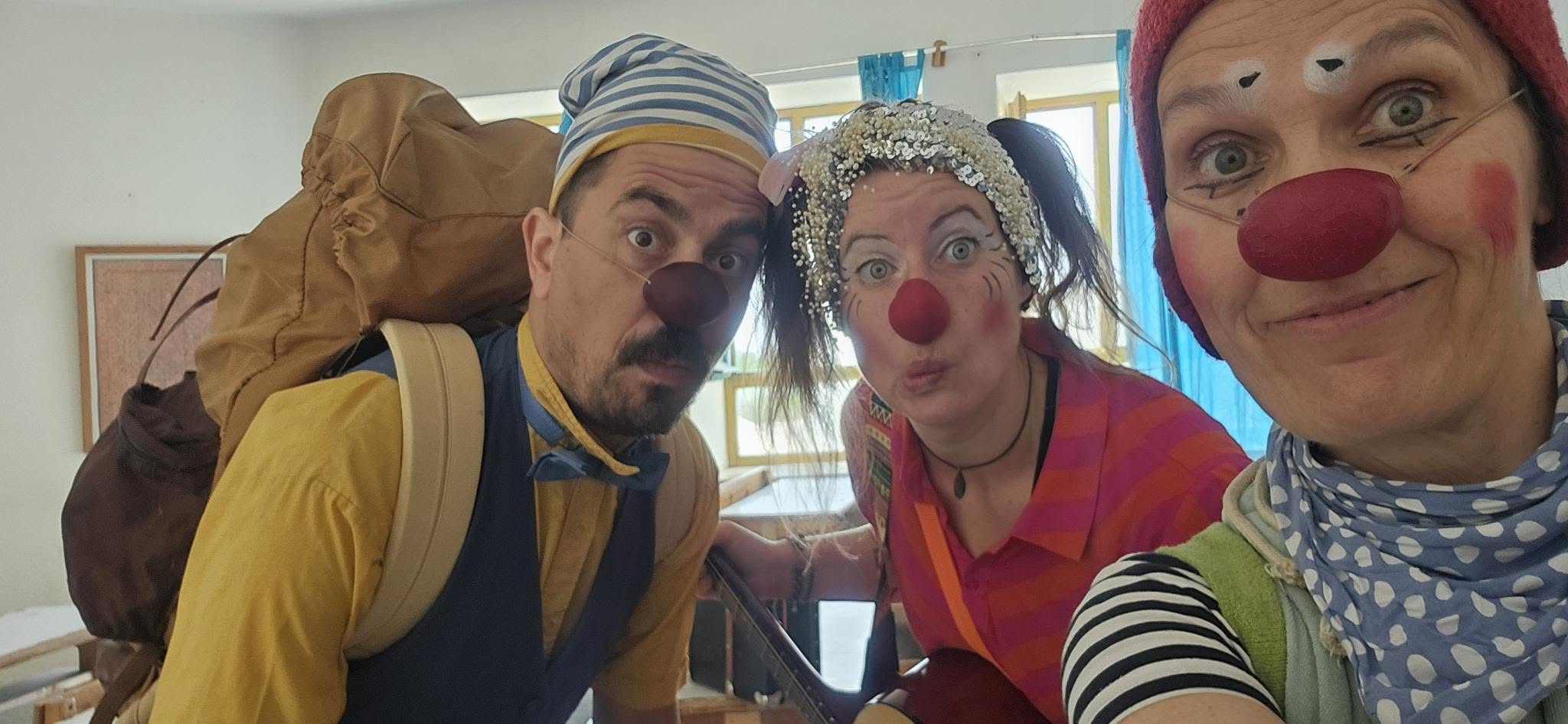 Drei Erwachsene, die als Clowns mit roten Nasen, Gesichtsbemalung und bunten Kostümen verkleidet sind, stehen drinnen und posieren für ein Selfie, wobei sie lustige Gesichter schneiden. Ein Clown trägt einen Rucksack, ein anderer einen glitzernden Hut.