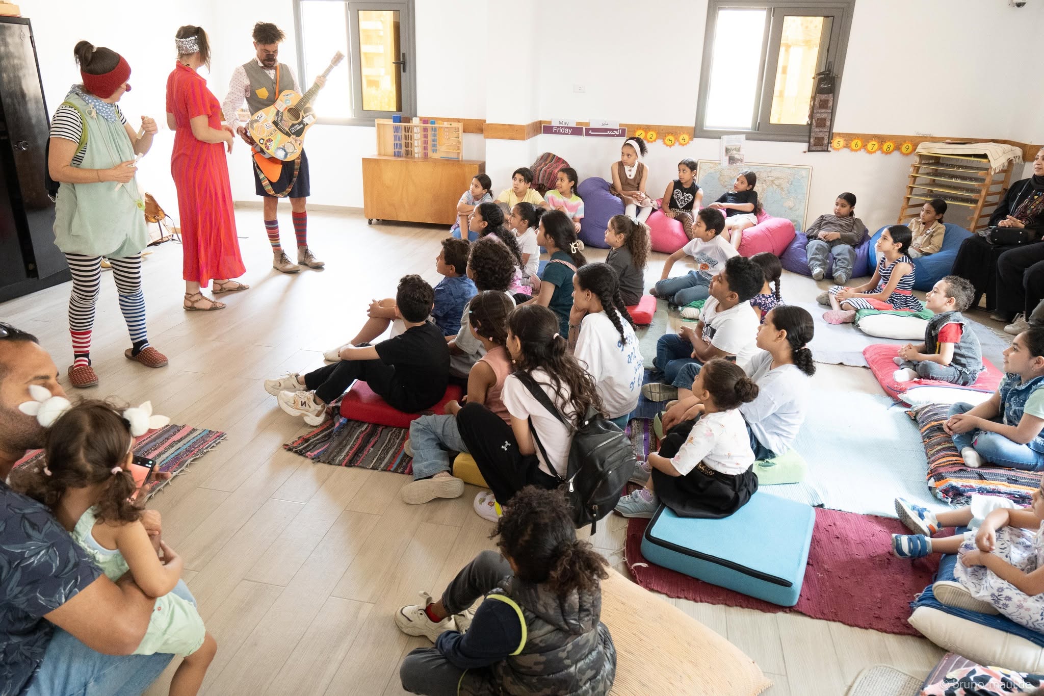 Eine Gruppe von Kindern sitzt auf Matten und Kissen in einem hellen Klassenzimmer und beobachtet aufmerksam zwei Darsteller in bunten Kostümen und mit bemalten Gesichtern, während die Erwachsenen an den Wänden sitzen.
