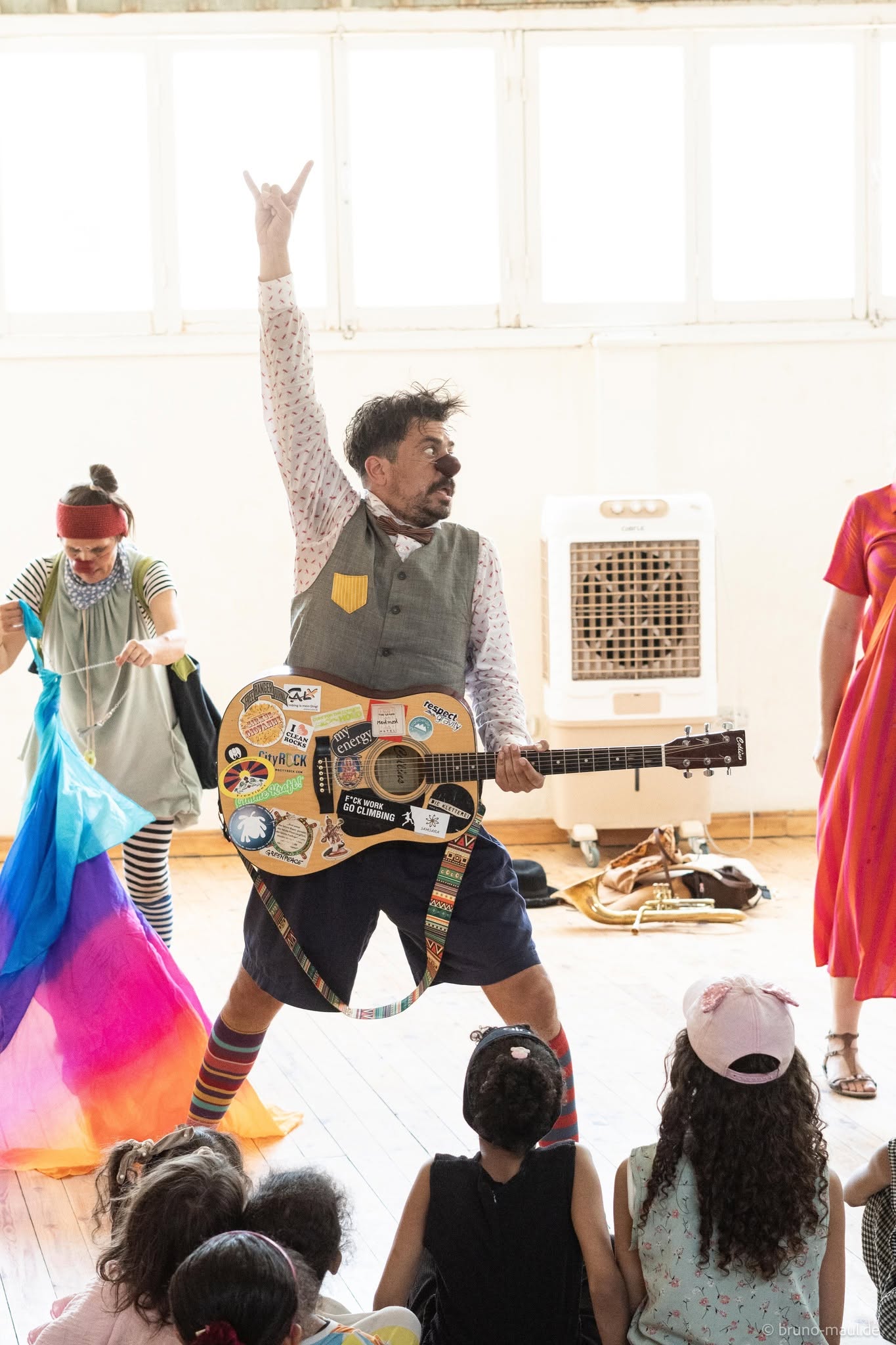 Ein Mann in bunten, clownartigen Kleidern spielt vor den Kindern voller Elan auf einer mit Aufklebern übersäten Gitarre. Weitere Darsteller in leuchtenden Kostümen stehen auf der Bühne und unterhalten die Kinder, die in einem hell erleuchteten Raum sitzen.