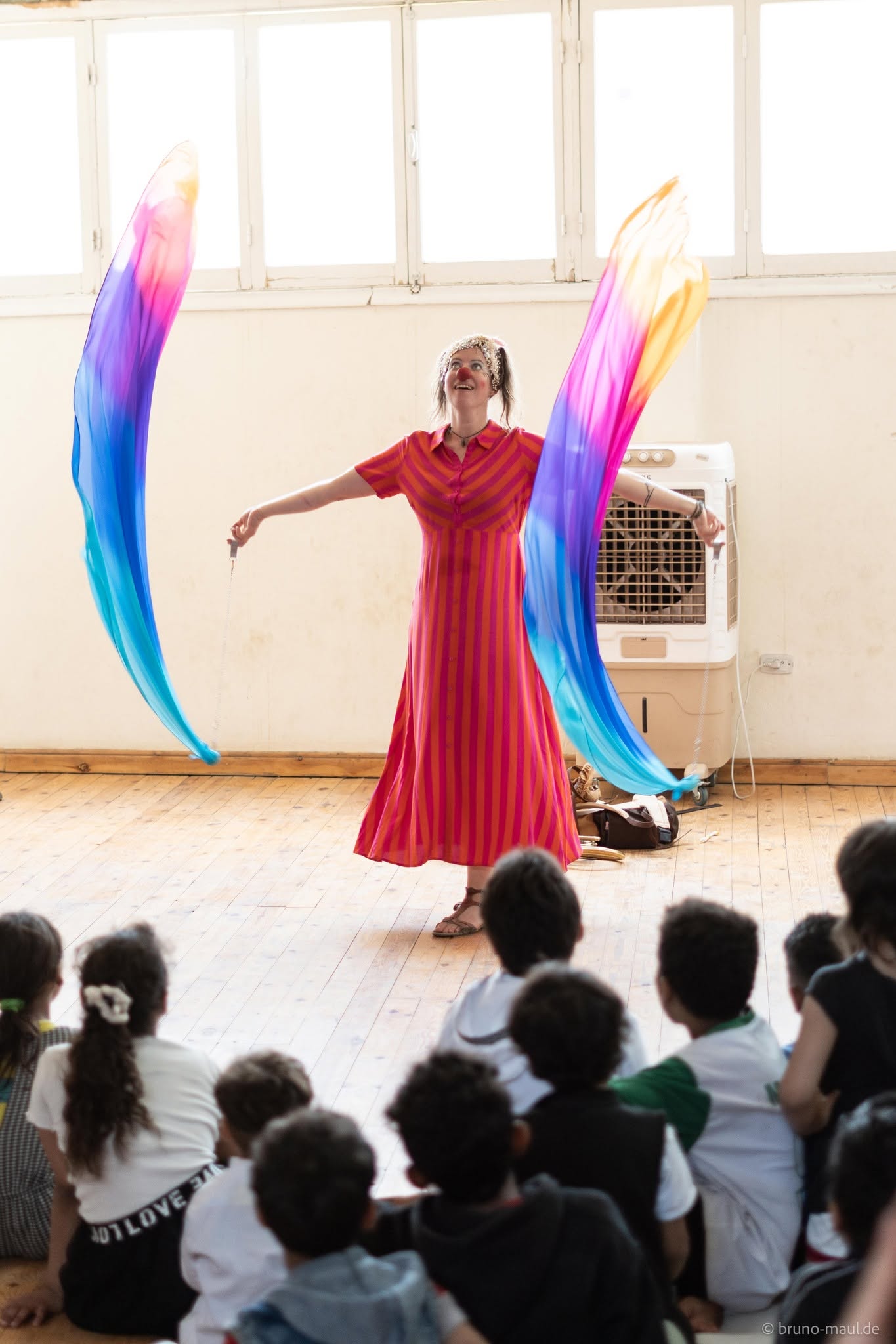 Eine Frau in einem leuchtend pink-rot gestreiften Kleid führt vor einer Gruppe sitzender Kinder in einem sonnendurchfluteten Raum mit Holzboden eine Darbietung mit bunten, fließenden Seidenfächern auf.