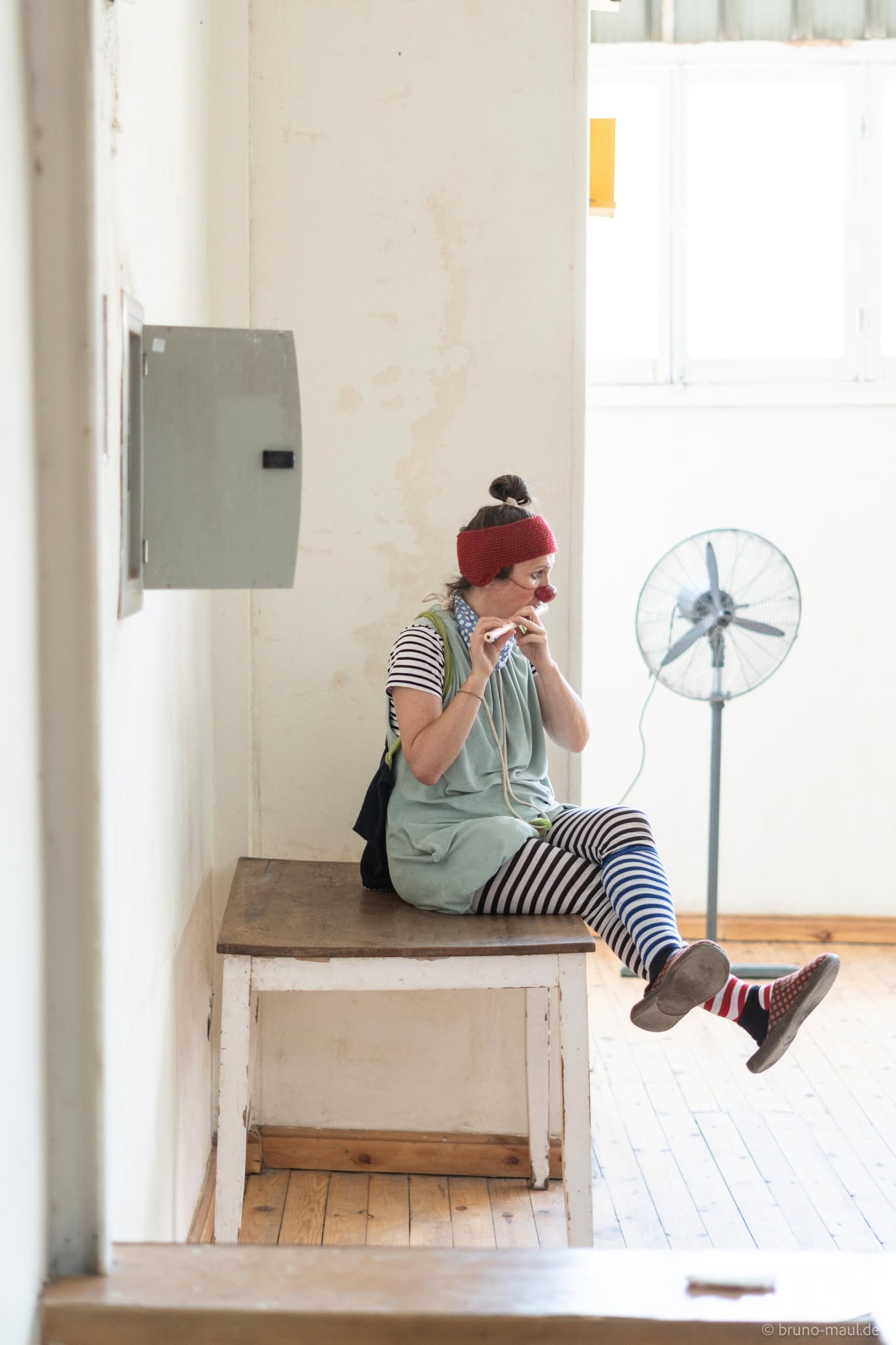 Eine als Clown verkleidete Person, die gestreifte Leggings und eine rote Mütze trägt, sitzt auf einer Bank an einer Wand und telefoniert mit einem Spielzeugtelefon. Im Hintergrund sind ein Standventilator und Sonnenlicht, das durch ein Fenster fällt, zu sehen.