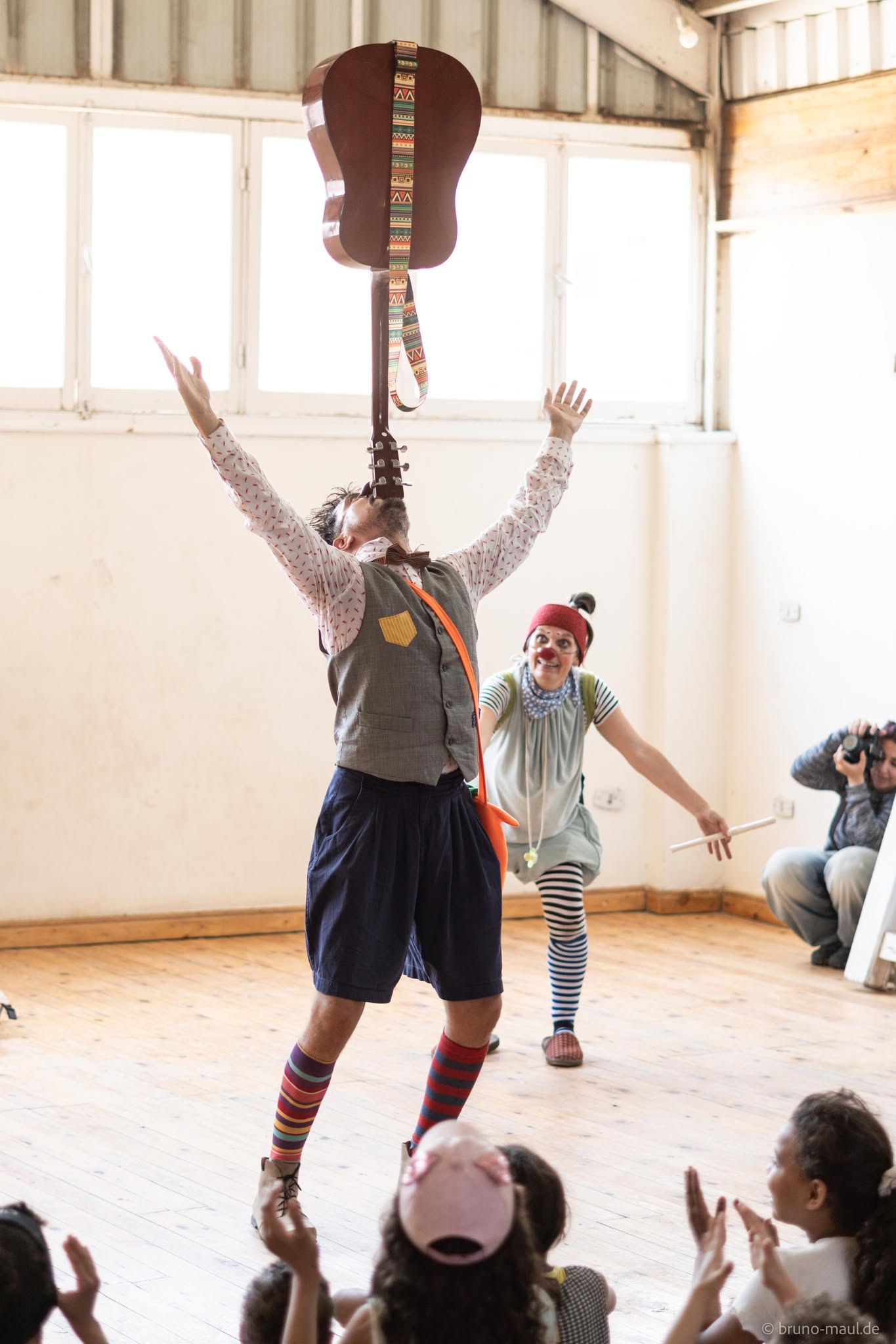 Ein Künstler balanciert eine Gitarre auf seinem Kinn, während er die Arme hochhält; hinter ihm steht ein Clown in bunten Kleidern und Socken. Vorne sitzen Kinder, die zuschauen und in dem sonnendurchfluteten Raum applaudieren.