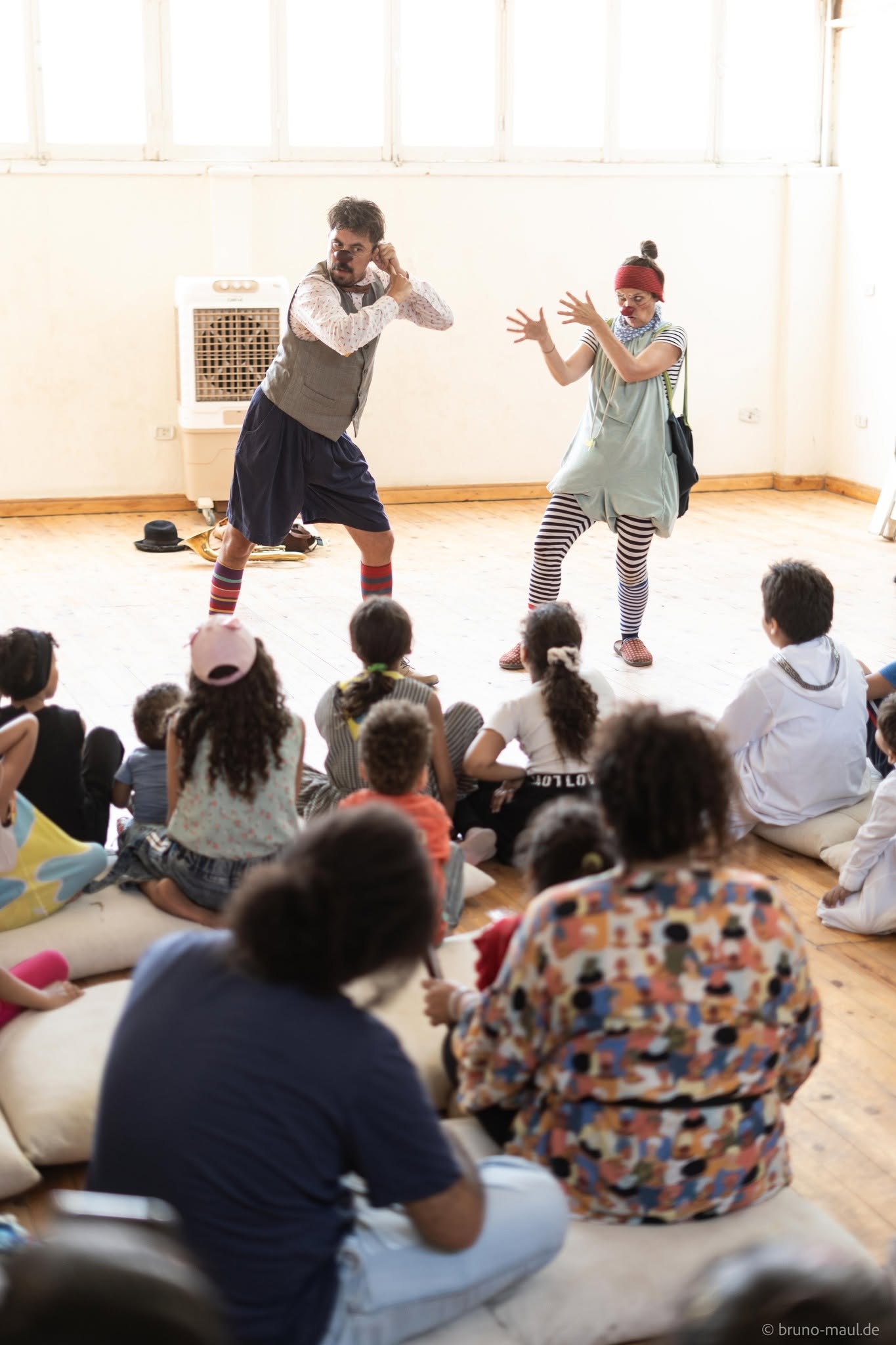 Zwei als Clowns verkleidete Darsteller unterhalten eine Gruppe von Kindern, die auf Kissen in einem hellen Raum mit Holzboden sitzen, mit ausdrucksstarken Gesten und verspielten Posen.