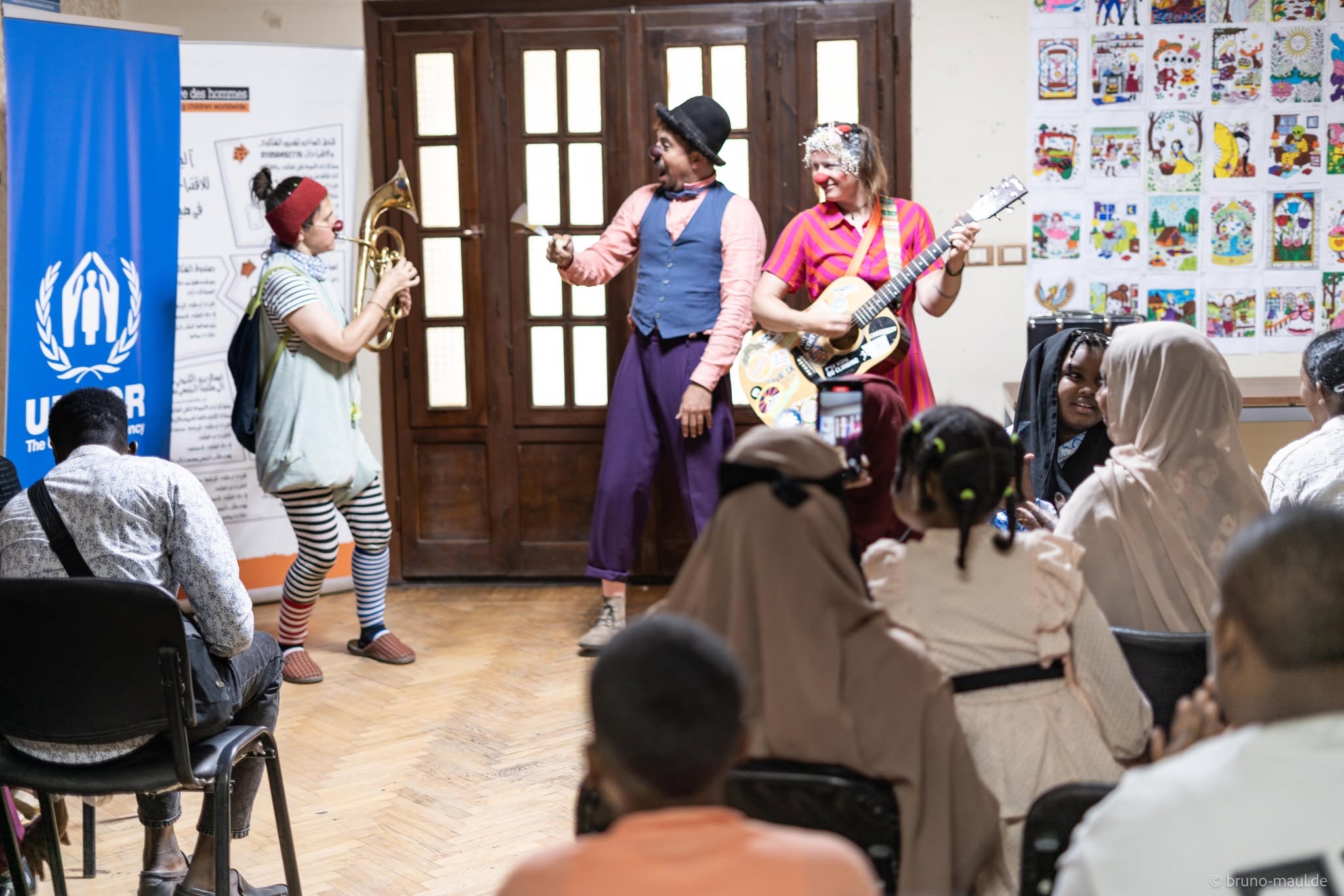Drei Clowns treten mit Musikinstrumenten vor einem Publikum aus Kindern und Erwachsenen auf. Plakate und Banner des UNHCR sowie bunte Zeichnungen schmücken den Hintergrund.