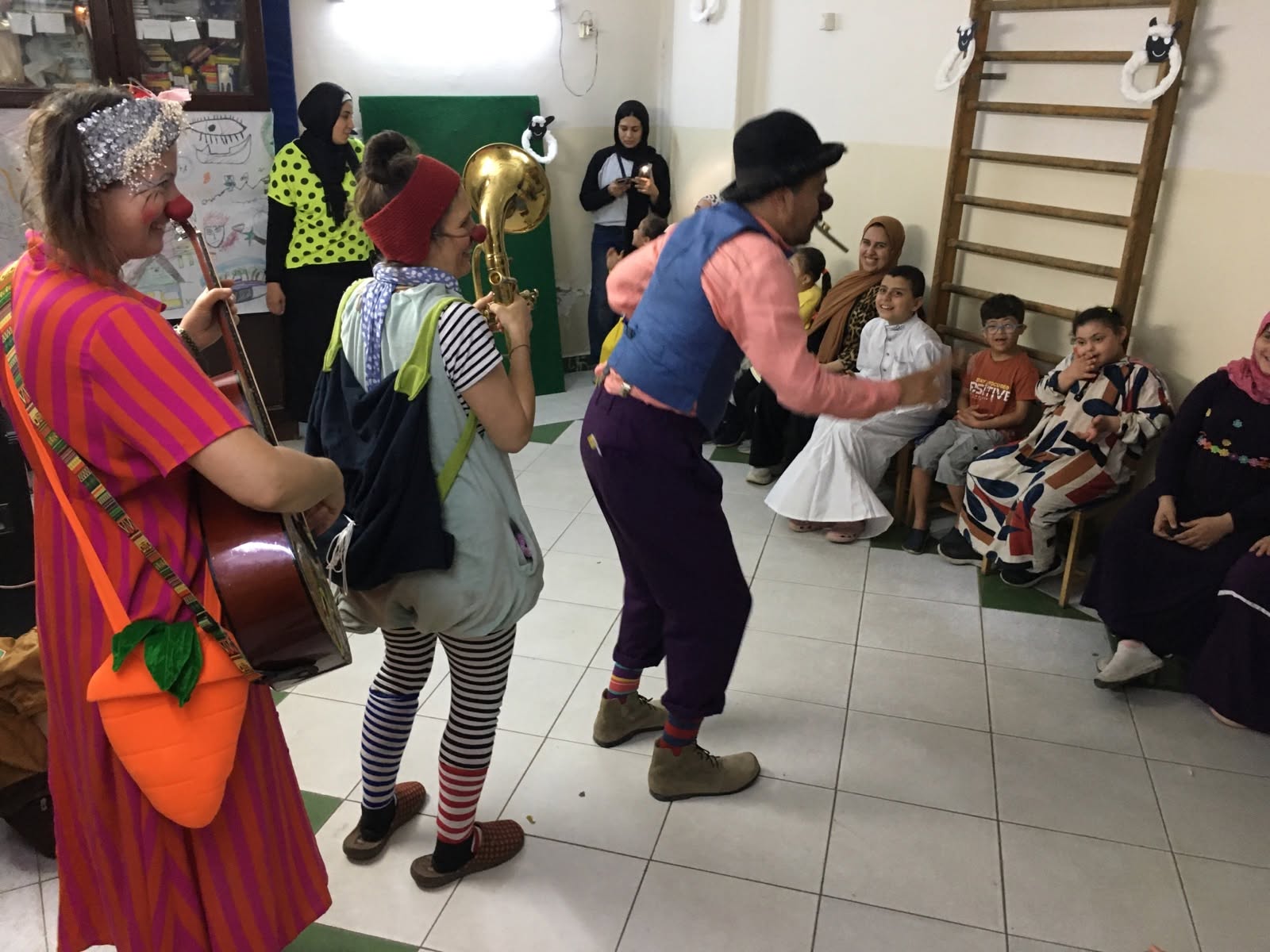 Drei Clowns in bunten Kostümen spielen Musik und führen vor einer Gruppe sitzender Kinder und Erwachsener in einem Raum eine Show auf, wodurch eine lebhafte und fröhliche Atmosphäre entsteht. Das Publikum lächelt und verfolgt die Vorstellung gebannt.