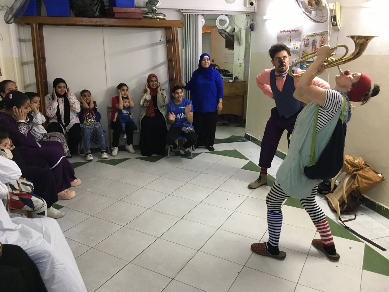 Zwei Clowns treten vor einer Gruppe von Kindern und Erwachsenen auf, die an den Wänden eines Raumes sitzen. Ein Clown spielt ein Blechblasinstrument, während der andere theatralisch gestikuliert. Das Publikum schaut interessiert und amüsiert zu.