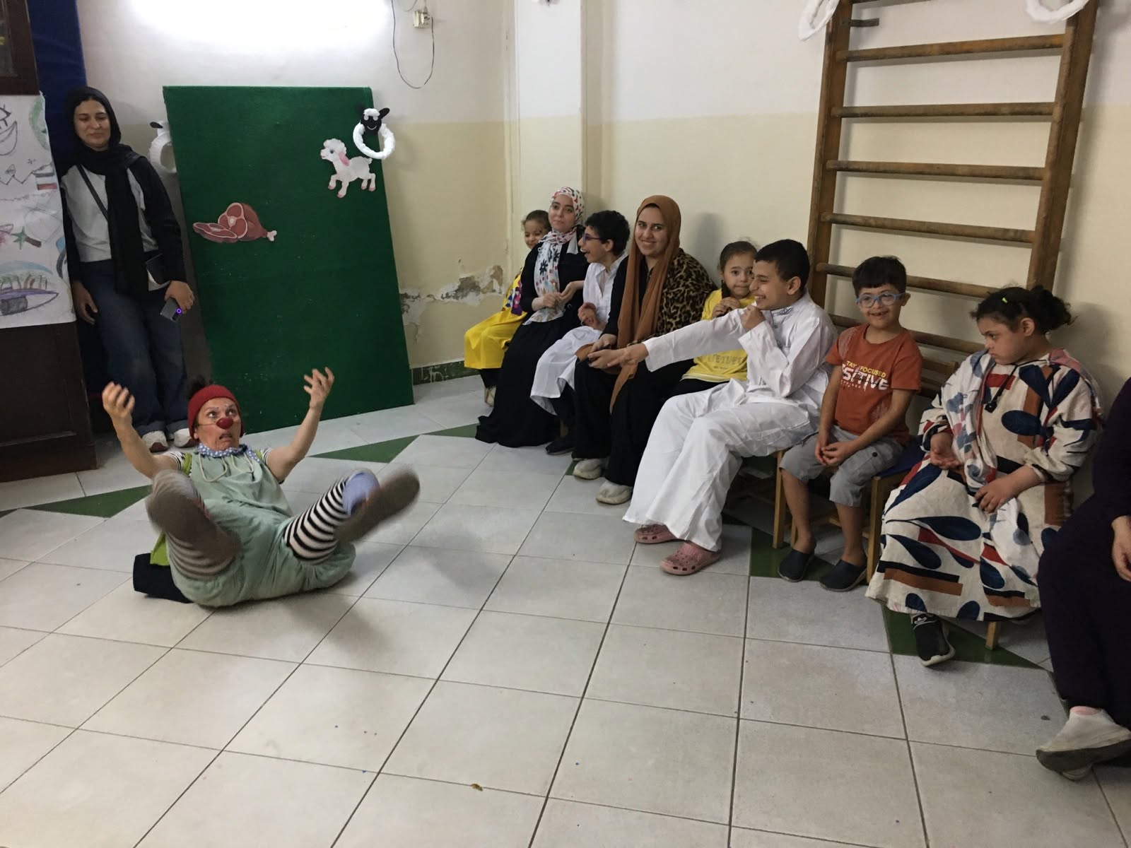 Ein Clown in einem grünen Kostüm mit gestreifter Hose fällt rückwärts um, während eine Gruppe von Kindern und Erwachsenen zusieht.