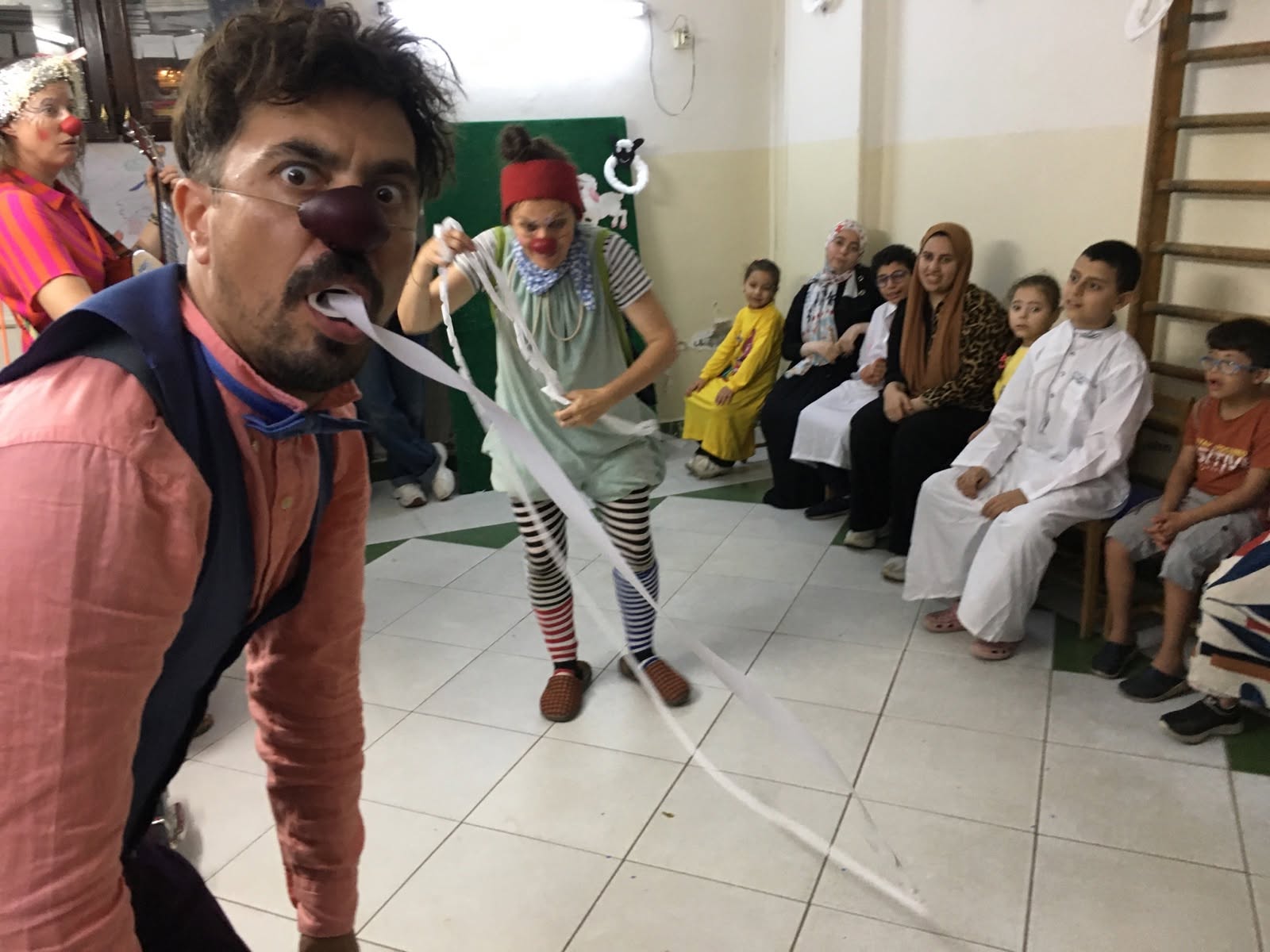 Ein männlicher Clown mit roter Nase und Fliege tut so, als würde er eine lange Luftschlange essen, während eine Clownin sie herauszieht. Mehrere Kinder und Erwachsene schauen zu und lächeln in einem hell erleuchteten Raum.