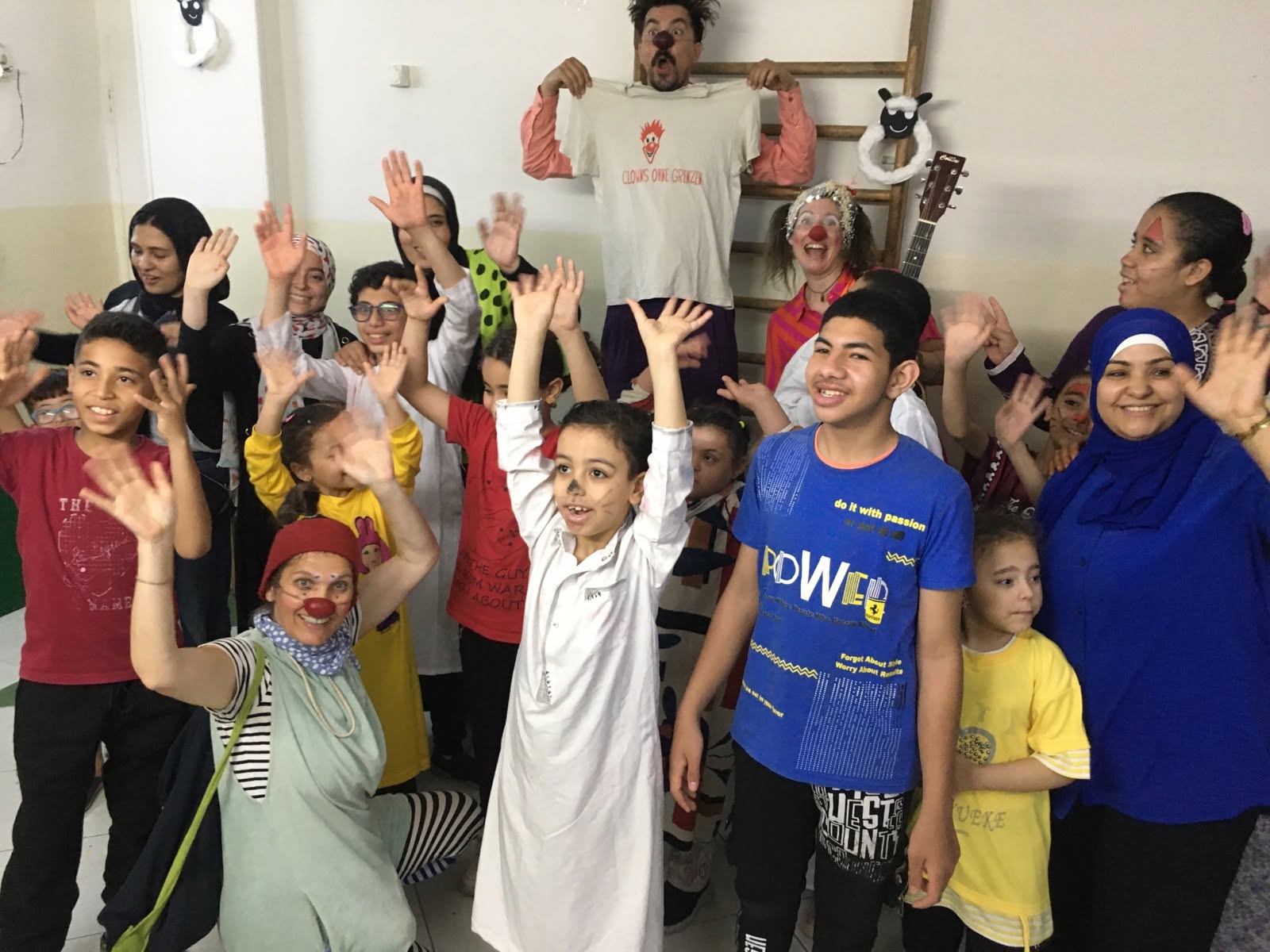 Eine Gruppe lächelnder Kinder und Erwachsener, von denen einige Kostüme tragen und Clownschminke im Gesicht haben, streckt in einem Raum fröhlich die Hände in die Höhe. Im Hintergrund hält jemand ein T-Shirt hoch, und an der Wand hängt eine Gitarre. Die Stimmung ist fröhlich und ausgelassen.