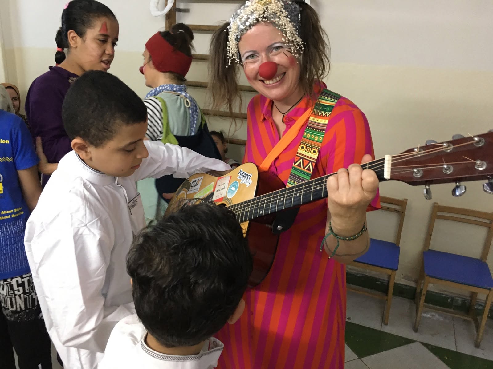 Ein lächelnder Clown mit roter Nase und buntem Kostüm spielt in einem Raum für Kinder Gitarre. Die Kinder sind ganz bei der Sache und schauen ihr zu. Im Hintergrund stehen mehrere Stühle an der Wand aufgereiht.